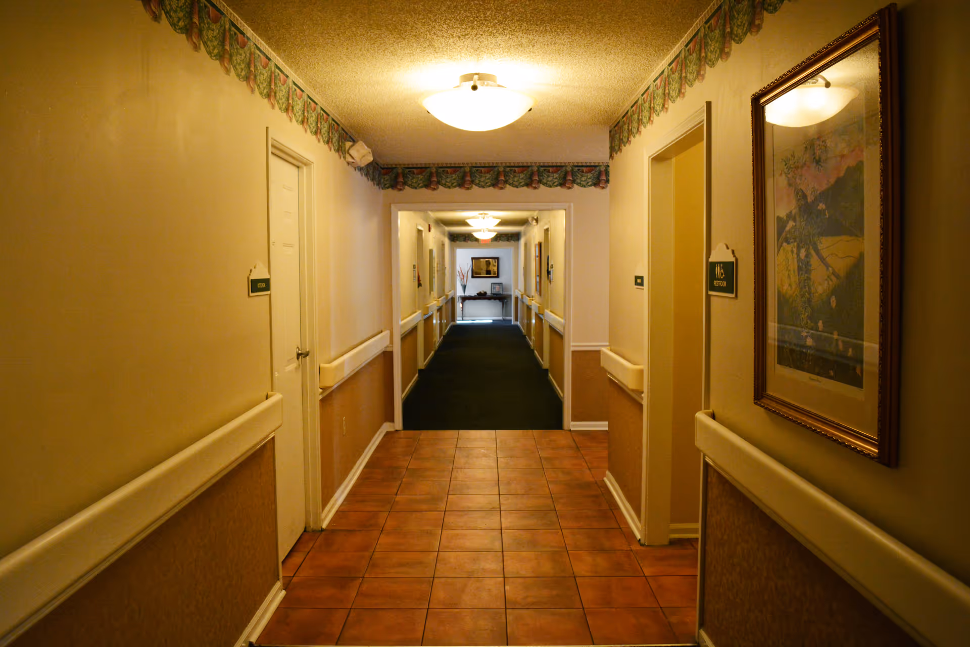 A well-lit hallway in a senior living facility with beige walls, tiled floor at the front transitioning to carpet further down the hall, handrails along both sides, several closed doors with room number signs, and a framed picture on the right wall.