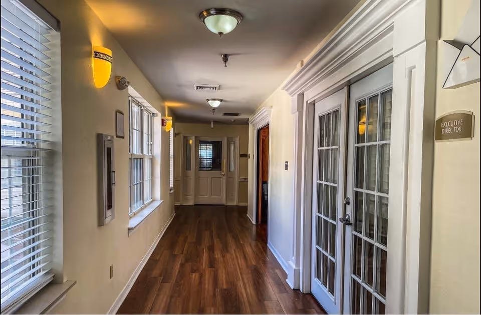 A well-lit hallway with wooden flooring and cream-colored walls. On the right side, there are double glass doors with a sign next to them that reads 'Executive Director.' The left side features several windows with blinds, and the hallway is illuminated by ceiling and wall-mounted lights.