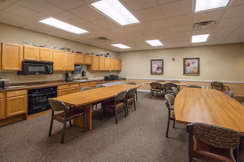 A spacious kitchen and dining area with wooden cabinets, a microwave, oven, and countertop appliances along one wall. Two long wooden tables with chairs are arranged in the center and to the side of the room. The walls are beige with two framed floral paintings hanging. The ceiling has recessed lighting and a drop ceiling design.