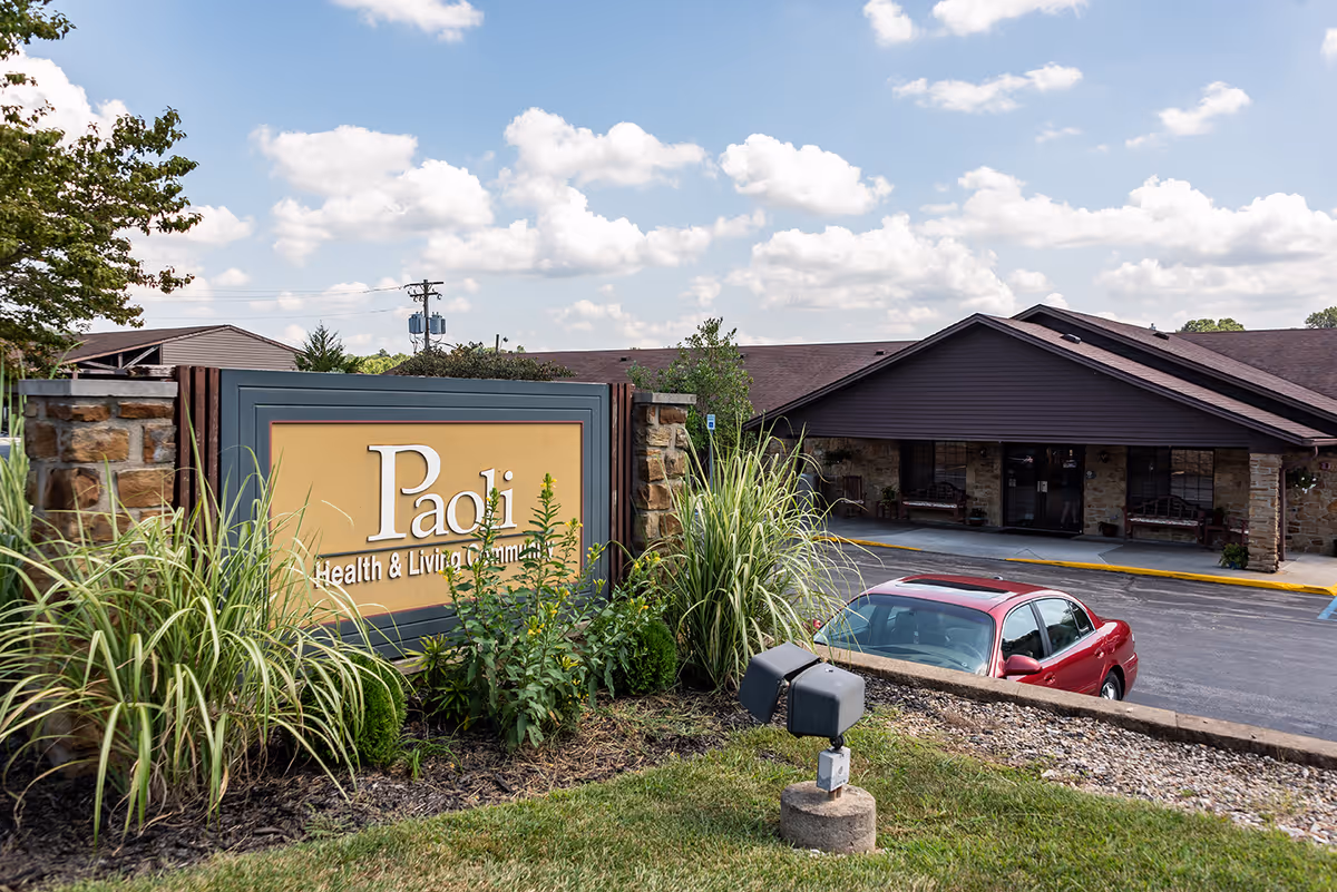 Exterior view of Paoli Health & Living Community building with a large sign in front surrounded by plants and a red car parked nearby under a partly cloudy sky.