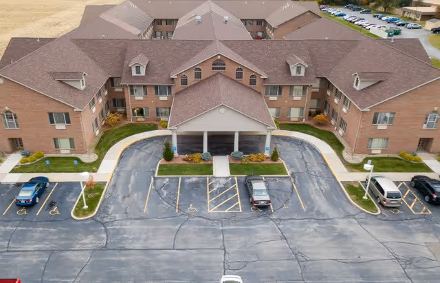 Aerial view of a large brick senior living facility with a covered entrance and multiple windows. The building has a brown roof and is surrounded by a parking lot with several cars and designated handicap parking spaces. There are sidewalks and small landscaped areas with grass and shrubs around the entrance.