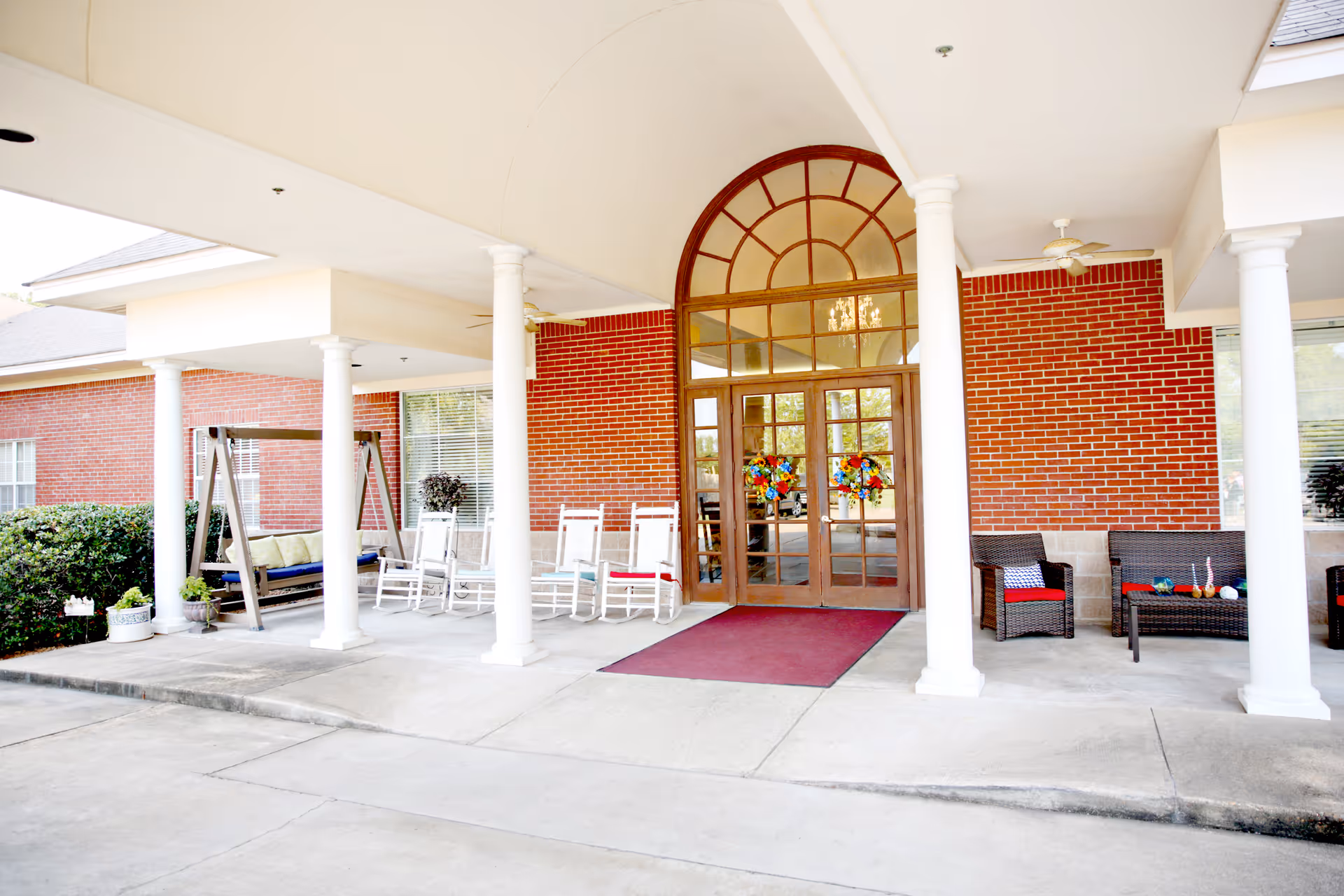 Entrance of a senior living facility with a covered porch supported by white columns. There are rocking chairs, a swing, and wicker seating with cushions arranged on the porch. The building has red brick walls and large glass double doors decorated with colorful wreaths. A red mat is placed in front of the doors.