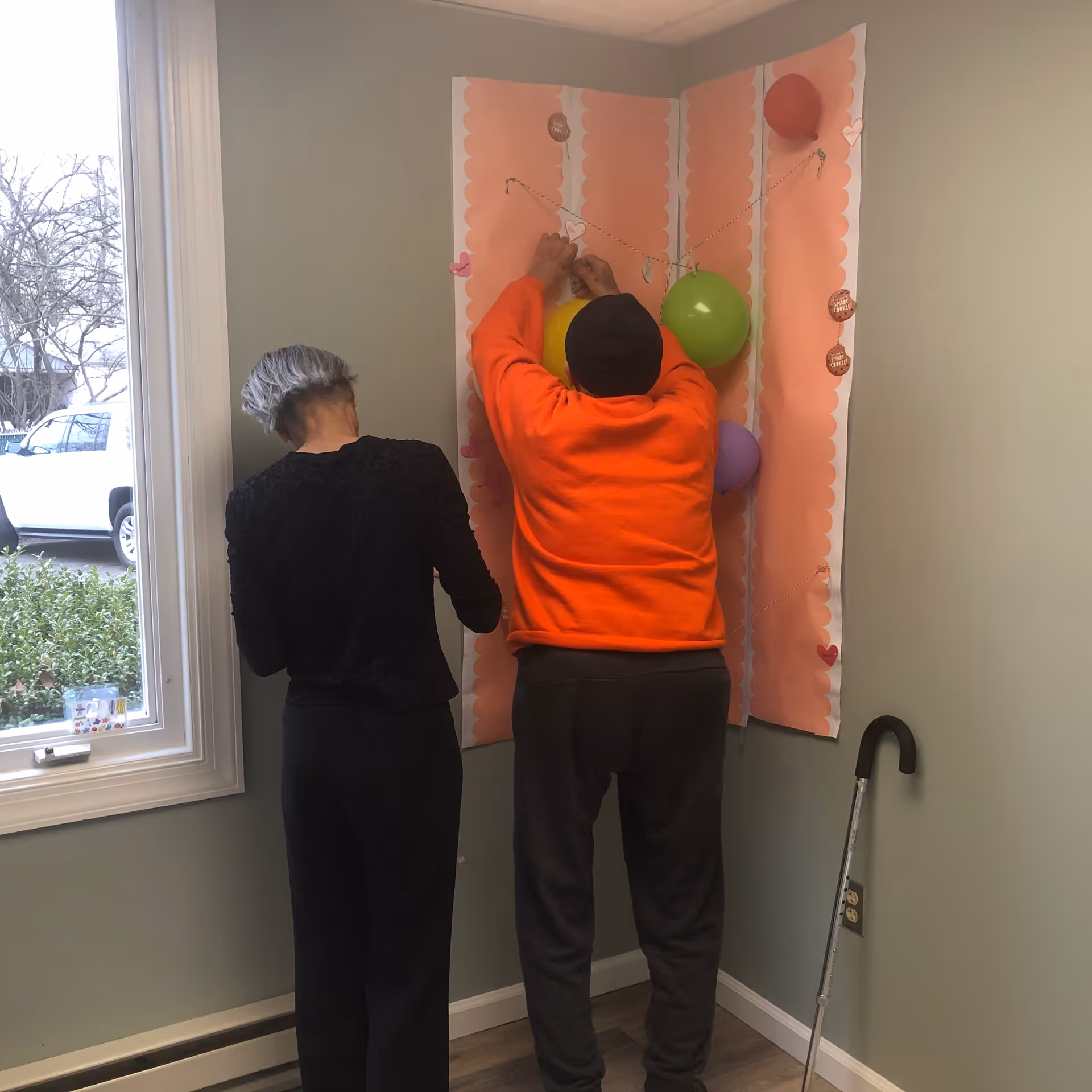 Two people decorating a corner wall with colorful balloons and paper cutouts. One person is wearing a bright orange sweatshirt and a black hat, while the other has gray hair and is dressed in black. A walking cane is leaning against the wall nearby, and a window shows a white vehicle and some greenery outside.