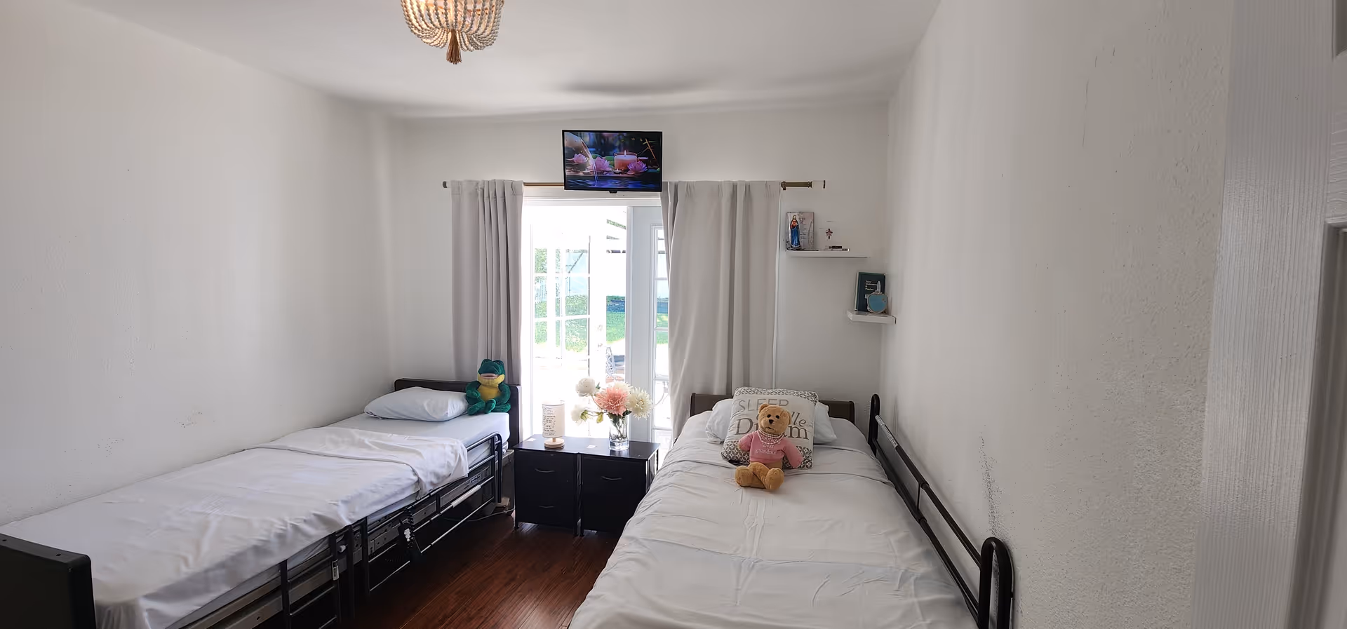 A small bedroom with two single beds placed against opposite walls. Each bed has white bedding; one bed has a green stuffed frog and the other has a teddy bear and a decorative pillow. Between the beds is a small black nightstand with flowers and other items on top. A window with white curtains is centered on the far wall, and a small TV is mounted above the window. The room has white walls and a wooden floor.