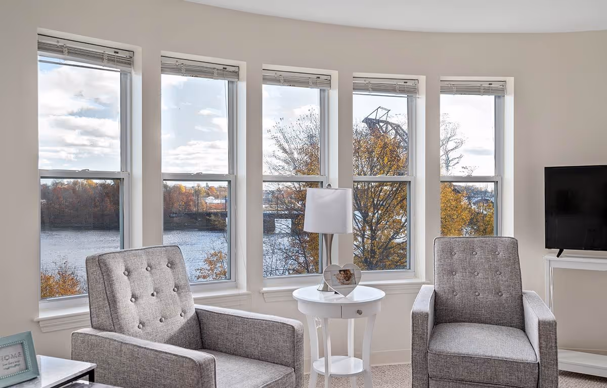 Two gray armchairs and a white side table with a lamp in front of large windows overlooking a river.