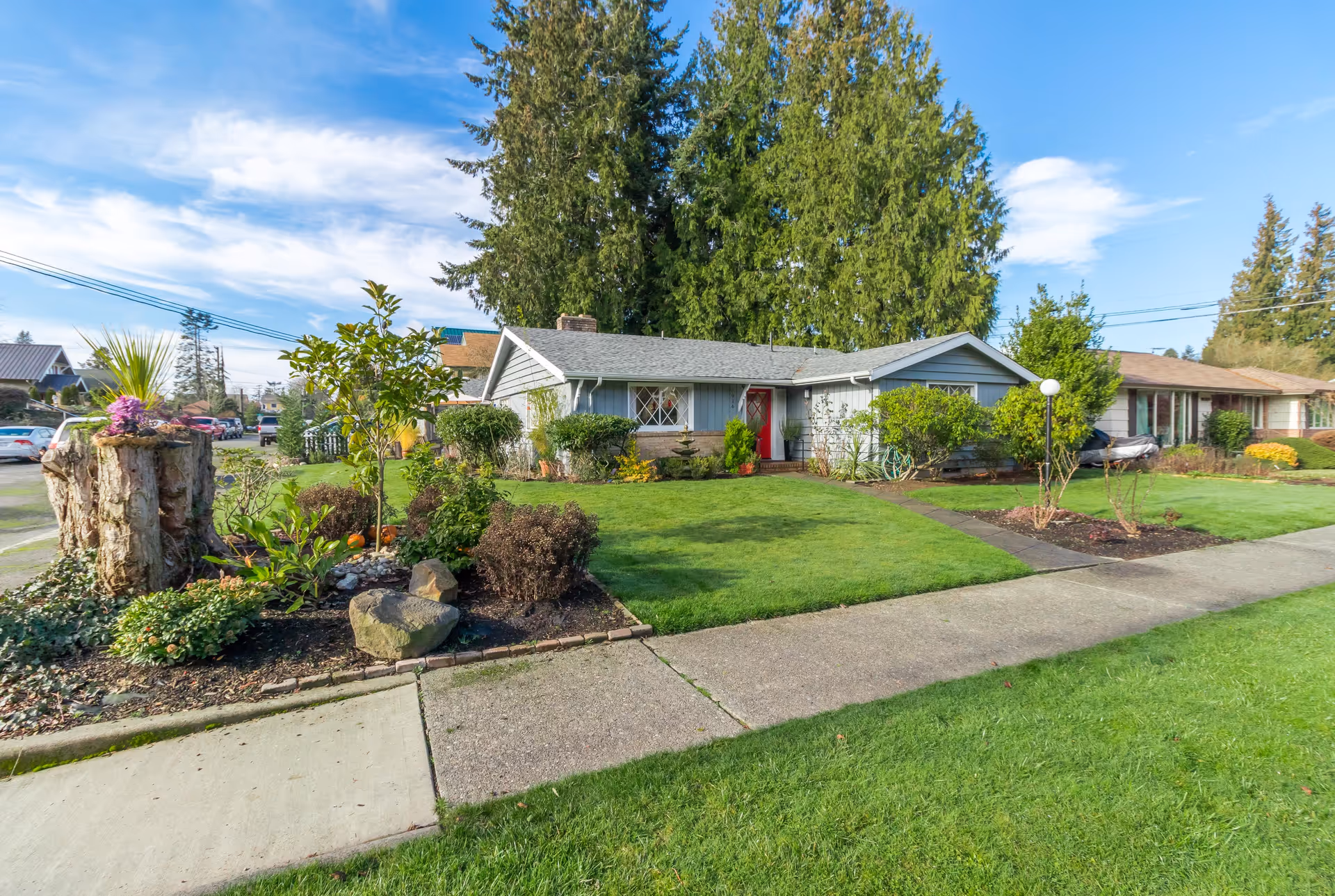 A single-story house with a gray exterior and a red front door, surrounded by a well-maintained lawn and garden with various shrubs and plants. There is a concrete sidewalk leading to the entrance and large trees in the background under a partly cloudy blue sky.