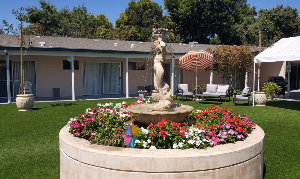 Outdoor courtyard area with a circular stone fountain featuring a statue in the center, surrounded by colorful flowers. In the background, there are patio chairs, a striped umbrella, potted plants, and a building with sliding glass doors and windows. Trees are visible behind the building under a clear blue sky.