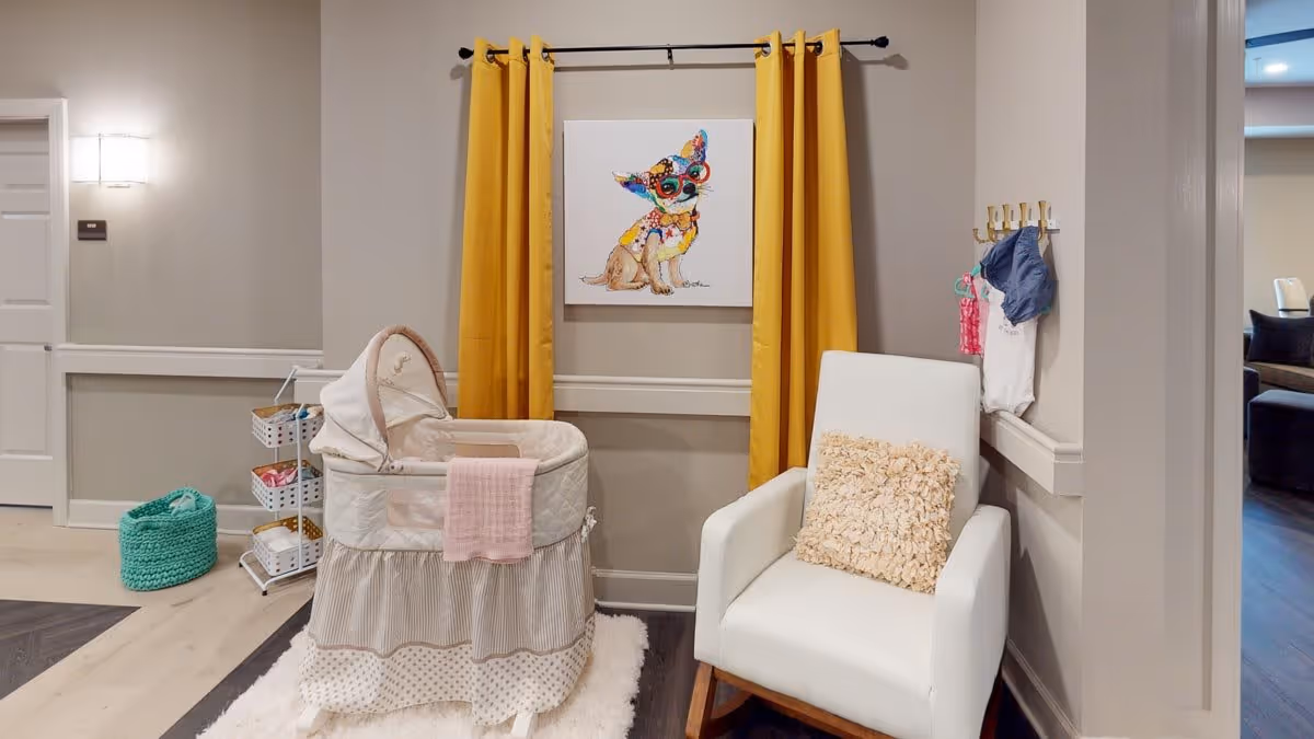 A cozy corner in a facility room featuring a white bassinet with a pink blanket draped over it, a white armchair with a textured beige pillow, yellow curtains framing a colorful dog painting on the wall, and a small rack with baskets and hanging baby clothes.