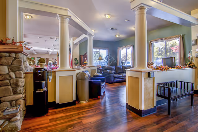 Interior view of a senior living facility common area with wooden flooring, white columns, a stone fireplace on the left, a water dispenser, trash can, black leather couches, a wooden bench, and windows with blinds letting in natural light.