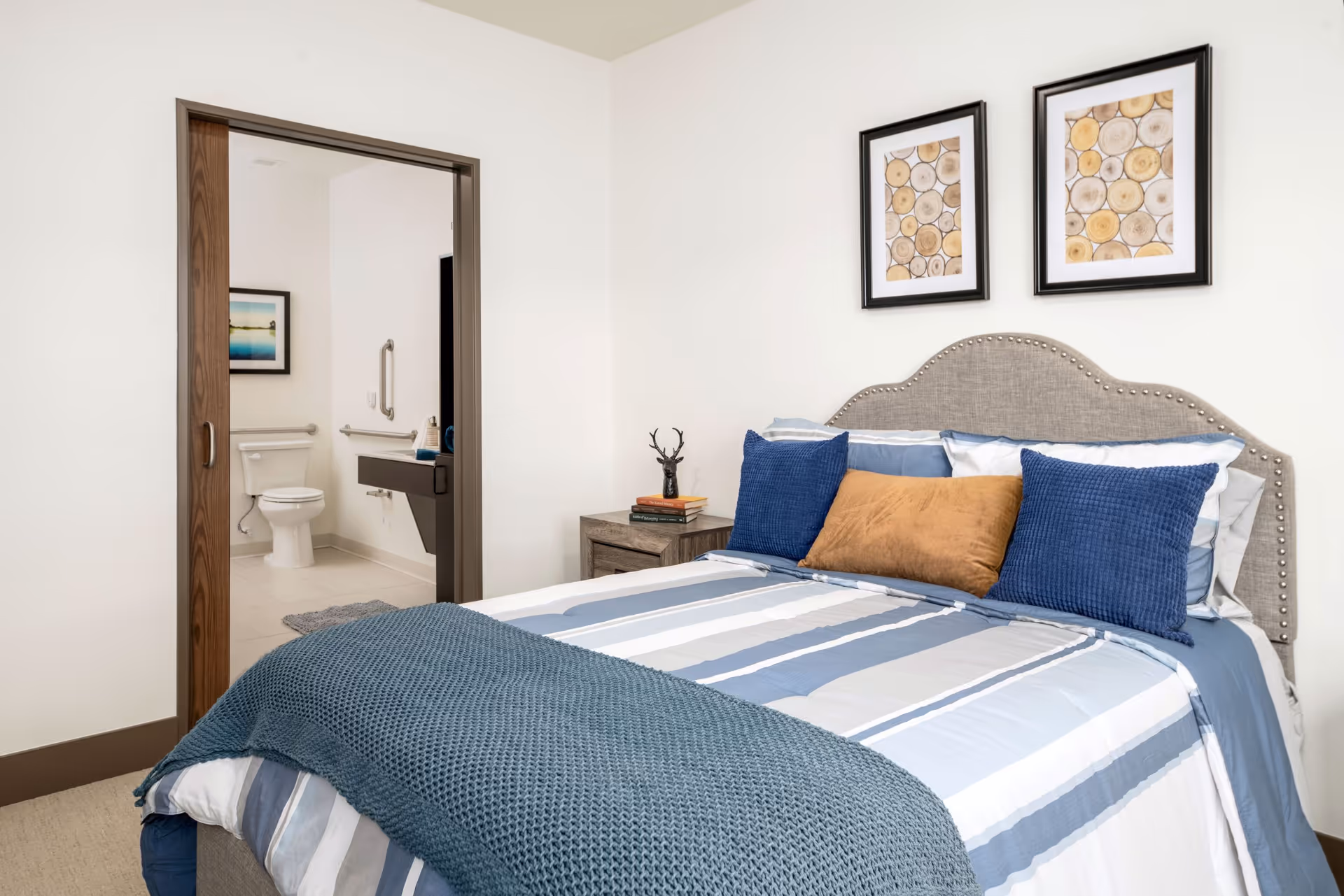 A neatly made bedroom with a striped bed, decorative pillows, a nightstand, and an open doorway revealing an accessible bathroom.