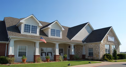 Front exterior of a suburban senior living building with a covered porch, rocking chairs, potted plants, and an American flag.