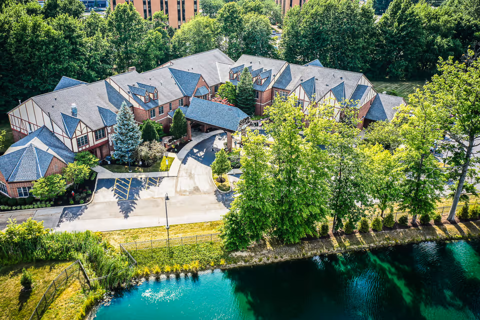 Aerial view of a large senior living facility building surrounded by trees and greenery, with a small pond in the foreground and a parking area in front of the building.