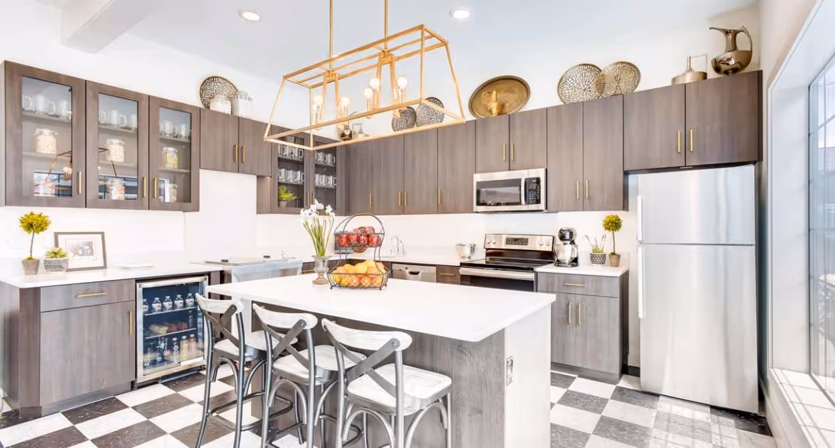 Bright modern kitchen with dark wood cabinets, white countertops, and a large island with three metal chairs. The kitchen features stainless steel appliances including a refrigerator, oven, microwave, and a small beverage fridge. Decorative items and plants are placed on the countertops and above the cabinets. The floor has a black and white checkered pattern, and a large window lets in natural light.