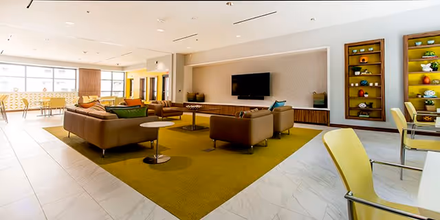 A spacious and well-lit living room area in a senior living facility featuring modern furniture including brown sofas and armchairs arranged around a central coffee table on a large mustard-colored rug. The room has a mounted flat-screen TV on the wall, decorative shelving units with various items, and large windows allowing natural light to fill the space. Additional seating with yellow chairs and tables is visible on the right side.