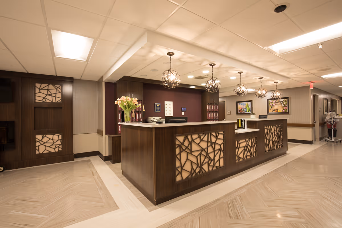 Reception area of a senior living facility with a wooden front desk featuring decorative paneling, modern pendant lights hanging from the ceiling, a vase of flowers on the counter, framed artwork on the walls, and a hallway leading to other rooms.