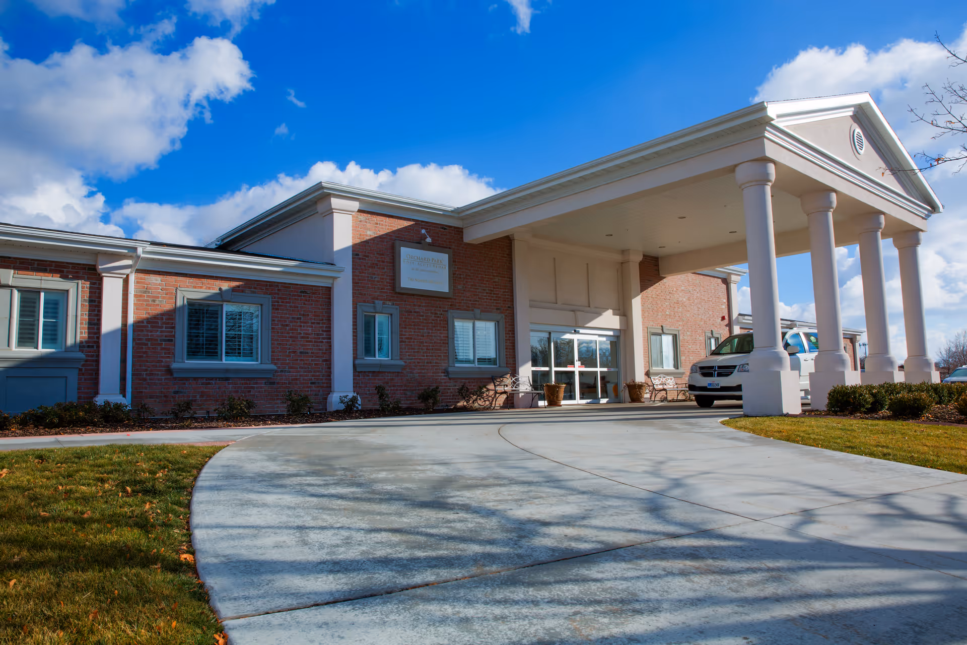 Front exterior view of a senior living facility named Cascades at Orchard Park, featuring a brick building with white columns supporting a covered entrance. A driveway curves up to the entrance, and a white vehicle is parked nearby under a bright blue sky with scattered clouds.