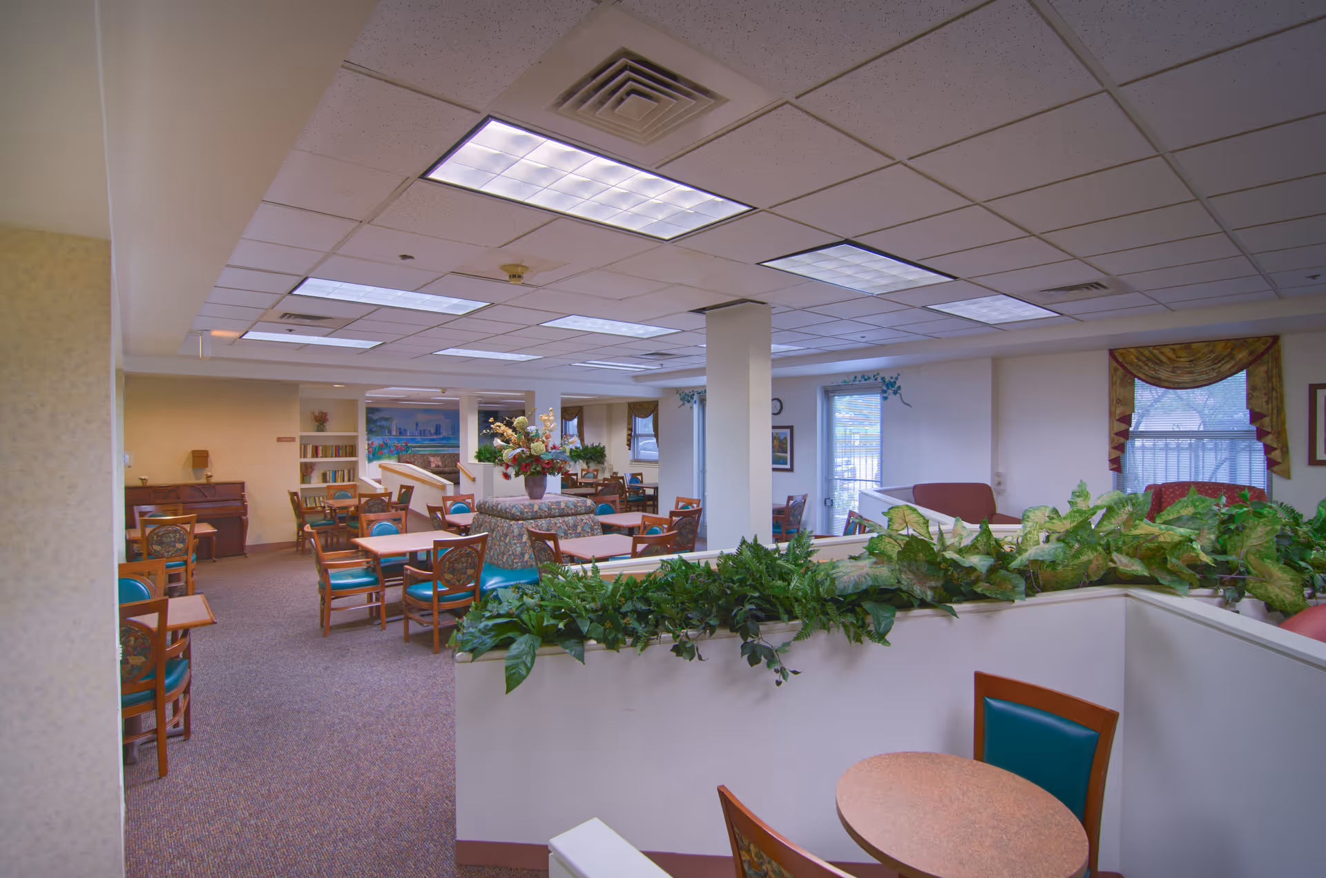 Interior view of a senior living facility common area with multiple tables and chairs arranged for dining or socializing. The room features carpeted floors, fluorescent ceiling lights, potted plants on a half wall, and windows with decorative curtains. There is a piano in the background and framed artwork on the walls.