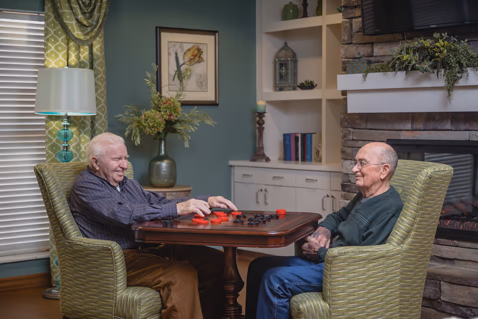 Two elderly men sitting in green armchairs playing checkers at a wooden table in a cozy room with a stone fireplace, built-in shelves, a lamp, and a window with green patterned curtains.