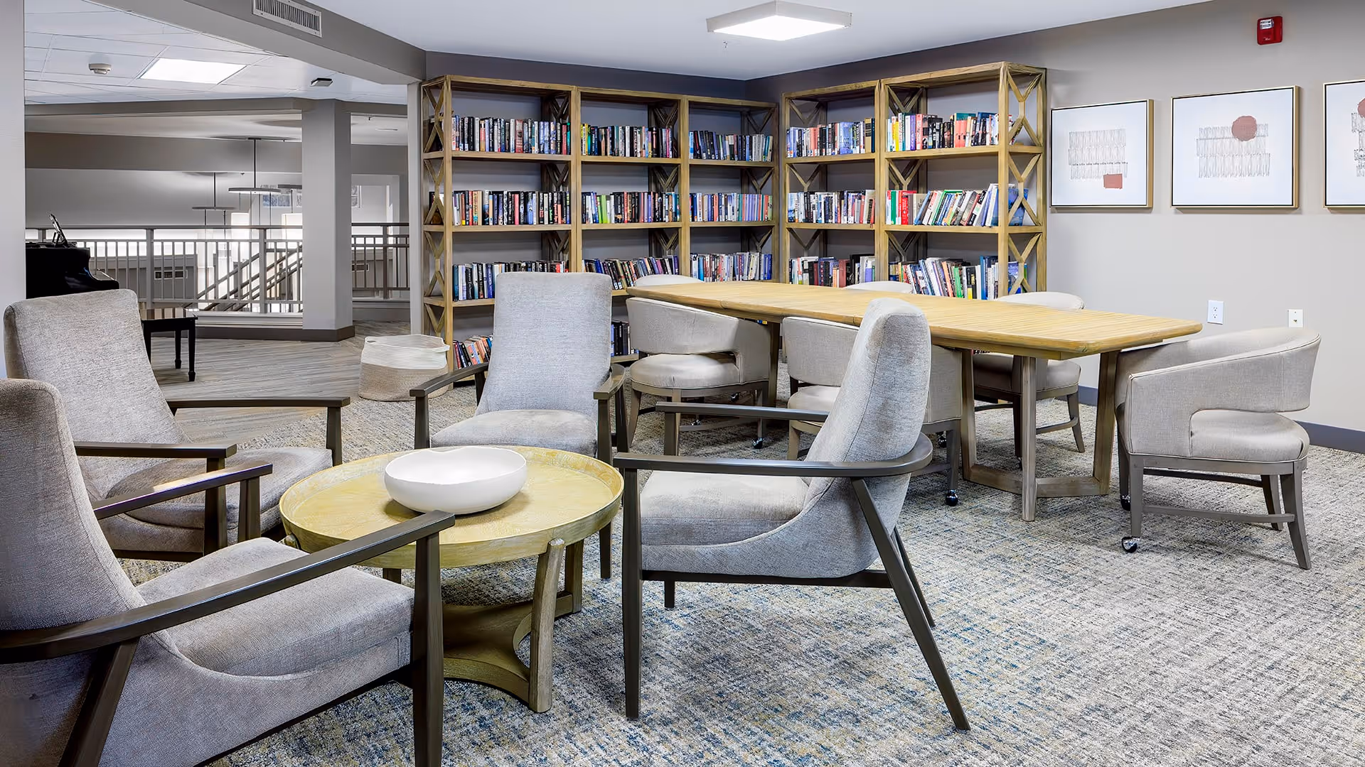 A cozy common area in a senior living facility featuring a seating arrangement with four cushioned armchairs around a round wooden table with a white bowl on it. Behind the seating area is a long rectangular wooden table surrounded by six cushioned chairs. In the background, there are bookshelves filled with books and three framed abstract artworks on the wall. The space has carpeted flooring and neutral-colored walls.