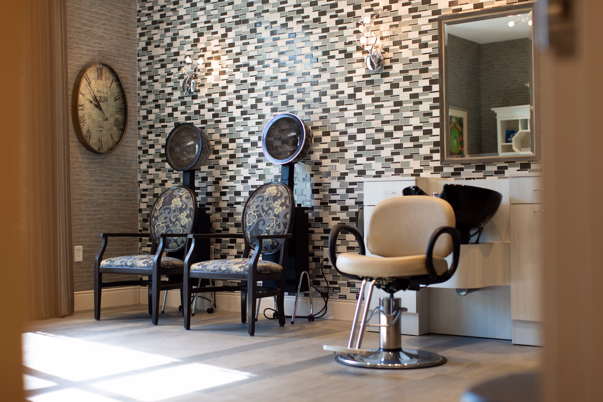 Interior salon area with two hooded hairdryers, two upholstered waiting chairs, a styling chair, sink, mirror and mosaic-tiled accent wall.