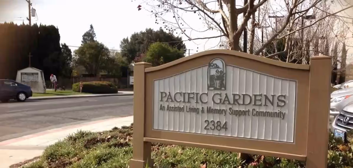A roadside entrance sign reading "Pacific Gardens — An Assisted Living & Memory Support Community, 2384" in front of landscaped grounds and parked cars.