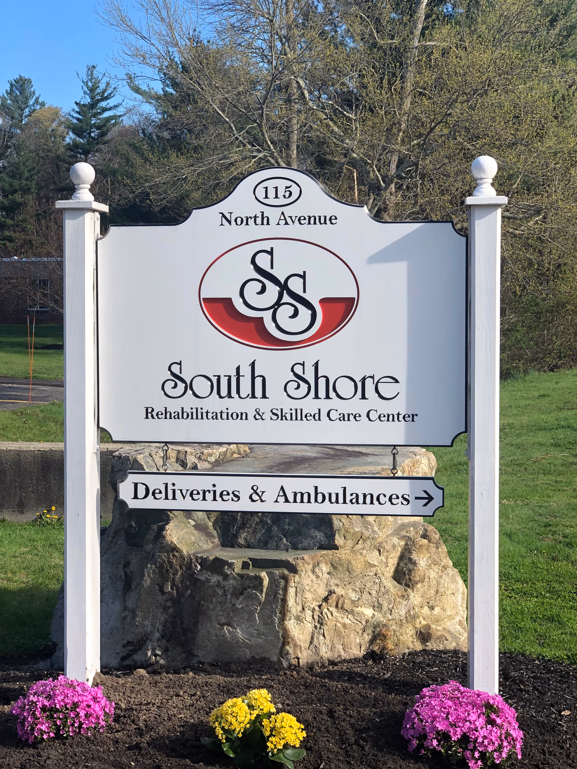 Entrance sign for South Shore Rehabilitation & Skilled Care Center at 115 North Avenue with a 'Deliveries & Ambulances' directional plaque in a landscaped grassy area with flowers.