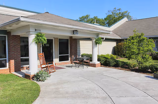 Exterior view of a single-story building with a covered entrance supported by white columns. There is a rocking chair and a small table with two chairs on the concrete patio. Green shrubs and a small tree are planted around the building, and the sky is clear and blue.