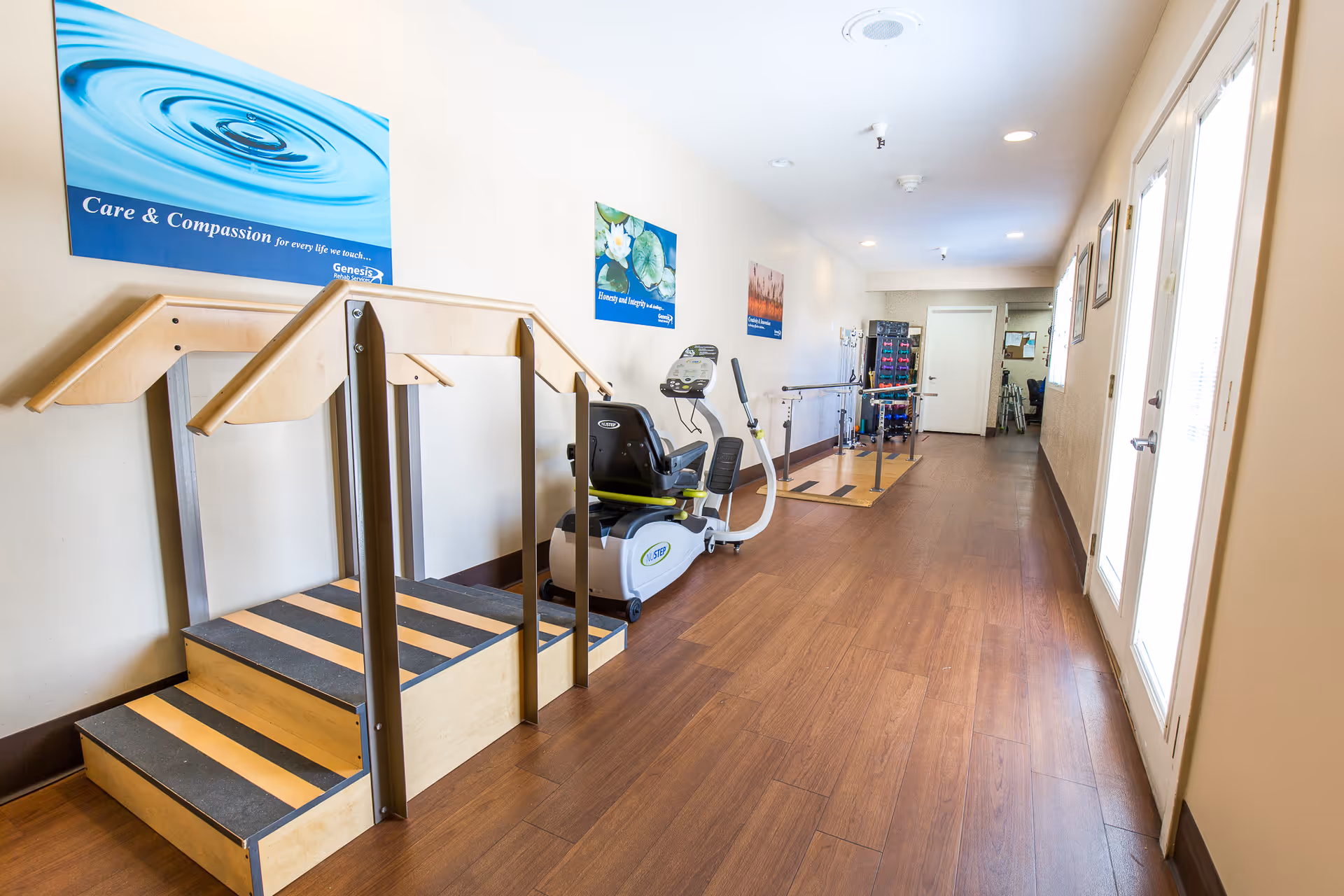 A bright rehabilitation hallway with wooden therapy steps, parallel bars, and an exercise bike along a long interior corridor.