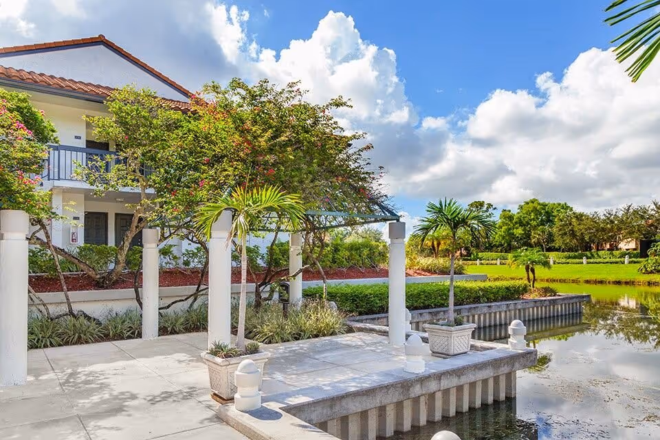 Lakeside patio with white columns, planters and a pergola in front of a two-story building beside a pond under a blue sky.