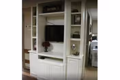 A white built-in entertainment center with shelves and cabinets. A flat-screen TV is mounted in the center, with decorative items such as a small plant and a 'HOME' sign on the top shelf. The surrounding area shows part of a hallway and other rooms in the background.