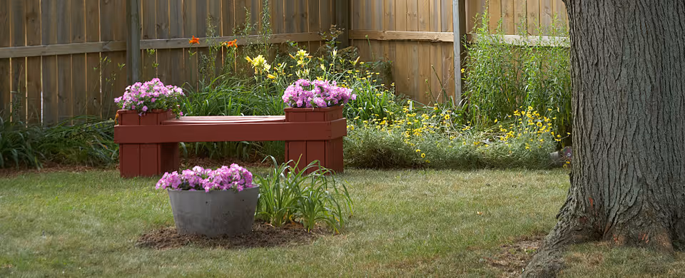 A garden area with a wooden fence in the background, a large tree trunk on the right, a red wooden bench with pink flowers on both ends, and a metal planter with pink flowers in the grass in the foreground. Yellow flowers and green plants are growing along the fence.