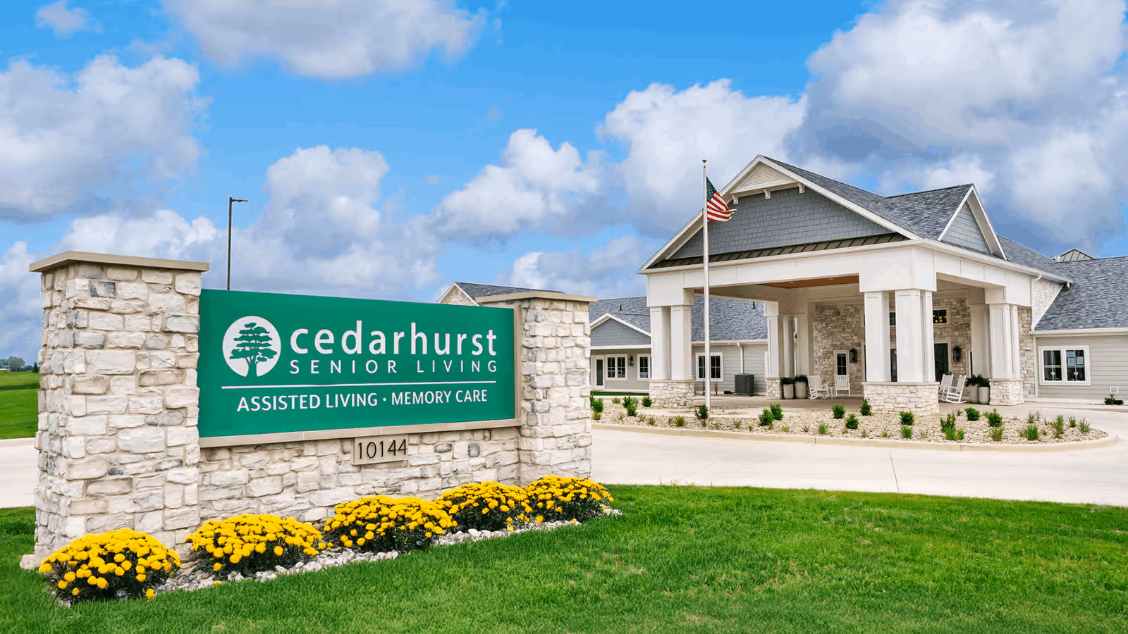 Exterior view of Cedarhurst Senior Living facility with a stone sign displaying the name and services offered, including assisted living and memory care. The building has a covered entrance with an American flag on a flagpole, surrounded by green grass and yellow flowers under a partly cloudy sky.