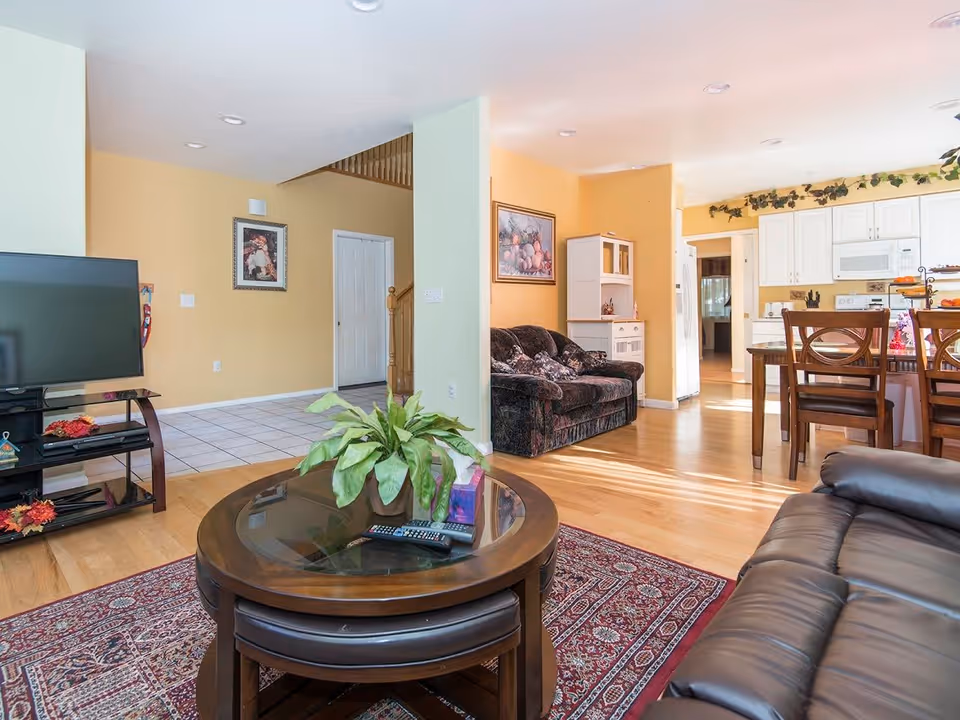 Interior view of a senior living facility showing a living room with a leather sofa, a round wooden coffee table with a plant and remote controls, a patterned rug, a TV on a stand, and an adjacent dining area with wooden chairs and a kitchen in the background. The walls are painted yellow and light green, and there are framed pictures and decorative plants.