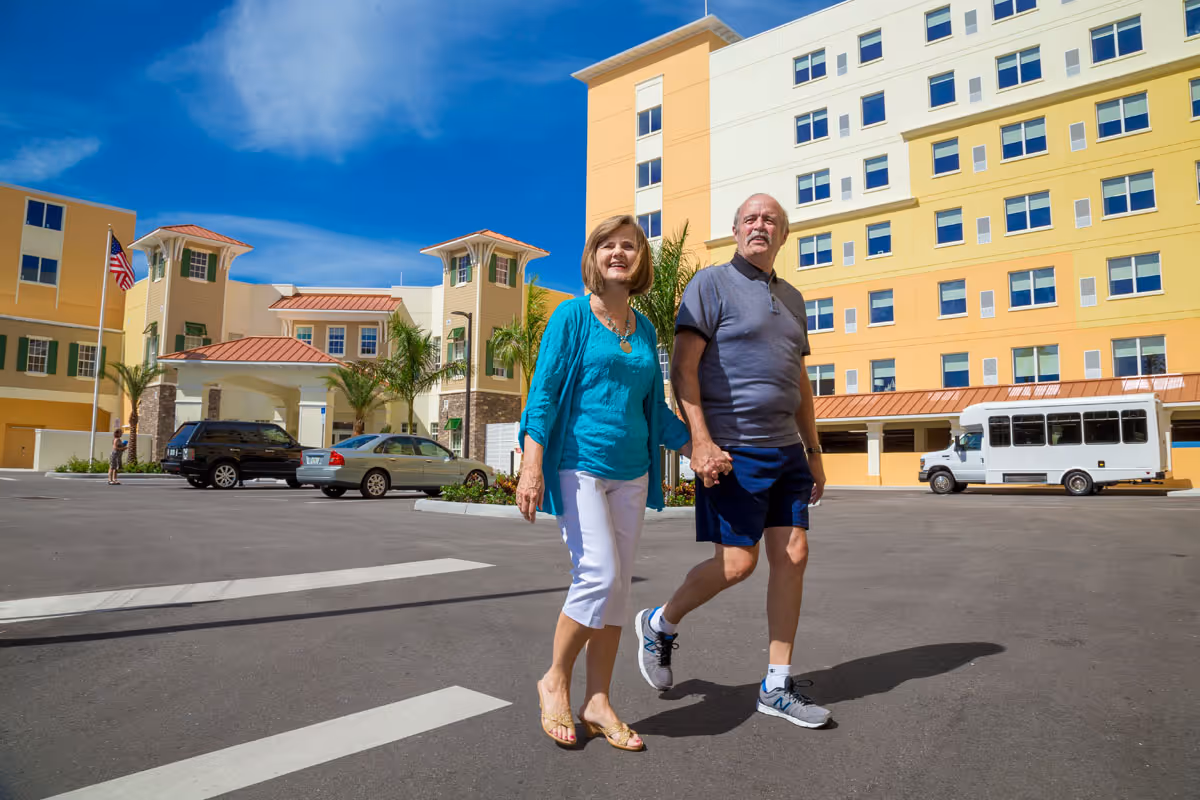 An elderly couple holding hands and walking in the parking lot outside a large, multi-story senior living facility with yellow and white exterior walls under a clear blue sky.