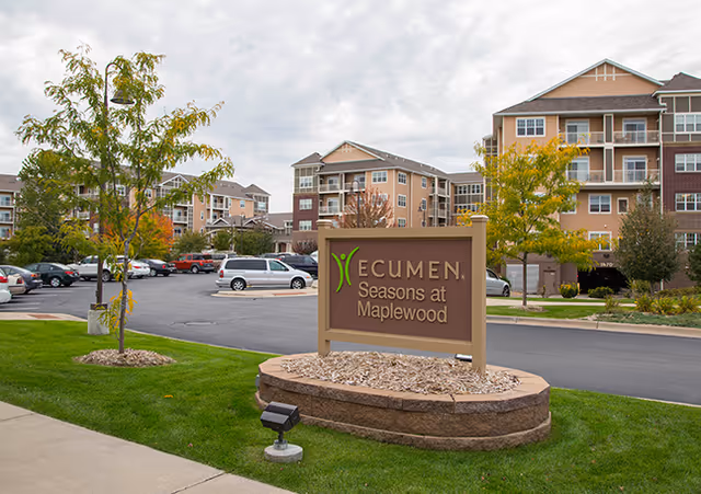 Exterior view of Ecumen Seasons at Maplewood senior living facility with a large sign in the foreground, multiple apartment buildings in the background, a parking lot with cars, and some trees with autumn foliage.