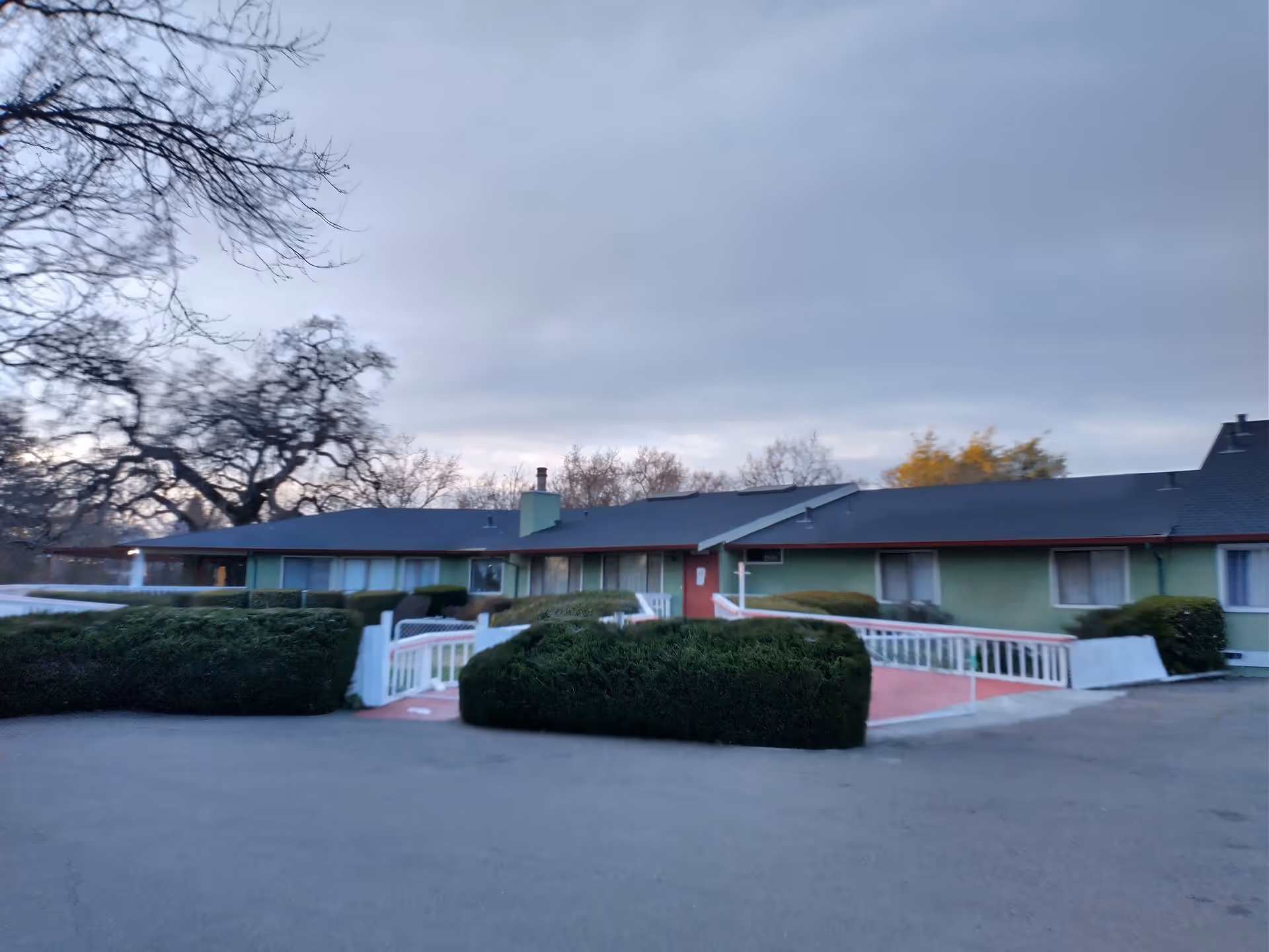 Exterior view of a single-story building with a dark roof and green walls, surrounded by trimmed bushes and leafless trees under a cloudy sky.