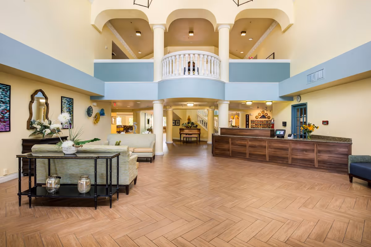 Spacious senior living facility lobby with light yellow walls and wood-patterned tile flooring. The area features a wooden reception desk on the right, a seating area with sofas and a table with decorative vases on the left, and a grand piano in the background. The upper level has a balcony with white balusters and columns supporting the ceiling with arches.