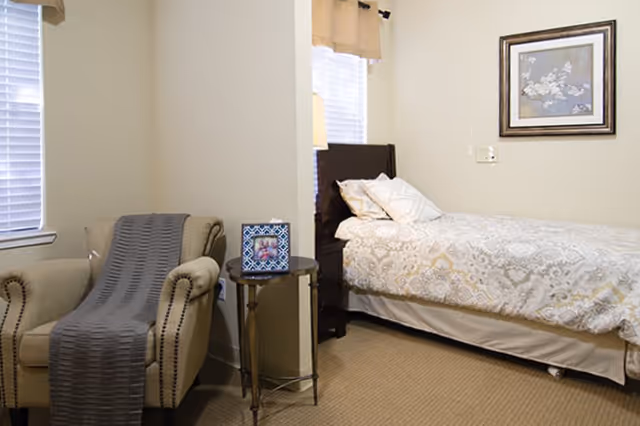 A tidy bedroom with a single bed dressed in patterned bedding, an upholstered armchair with a gray throw, a small round side table, and framed wall art.