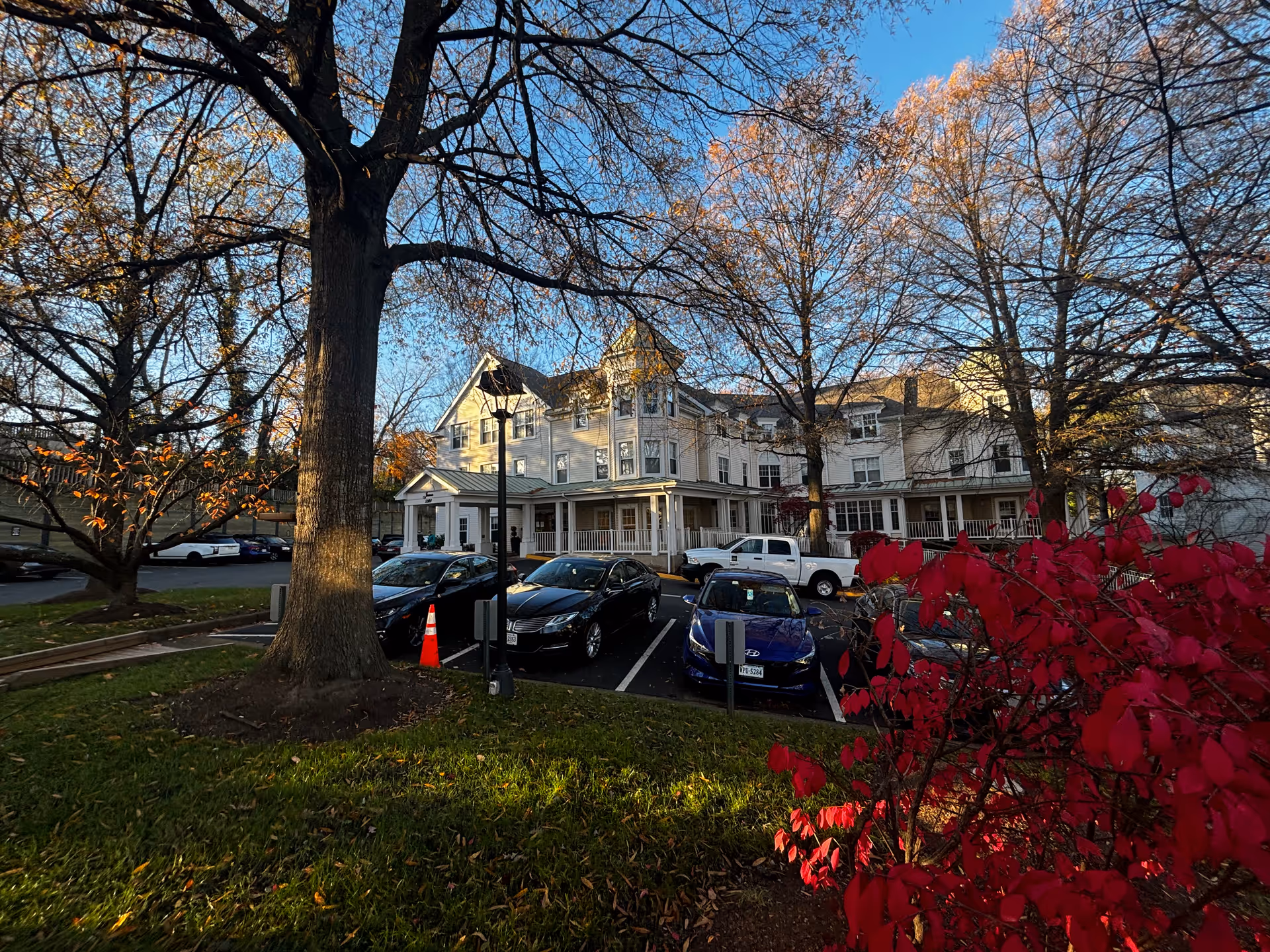 Front exterior of a multi-story senior living building framed by trees and autumn foliage with a parking lot of cars in front.