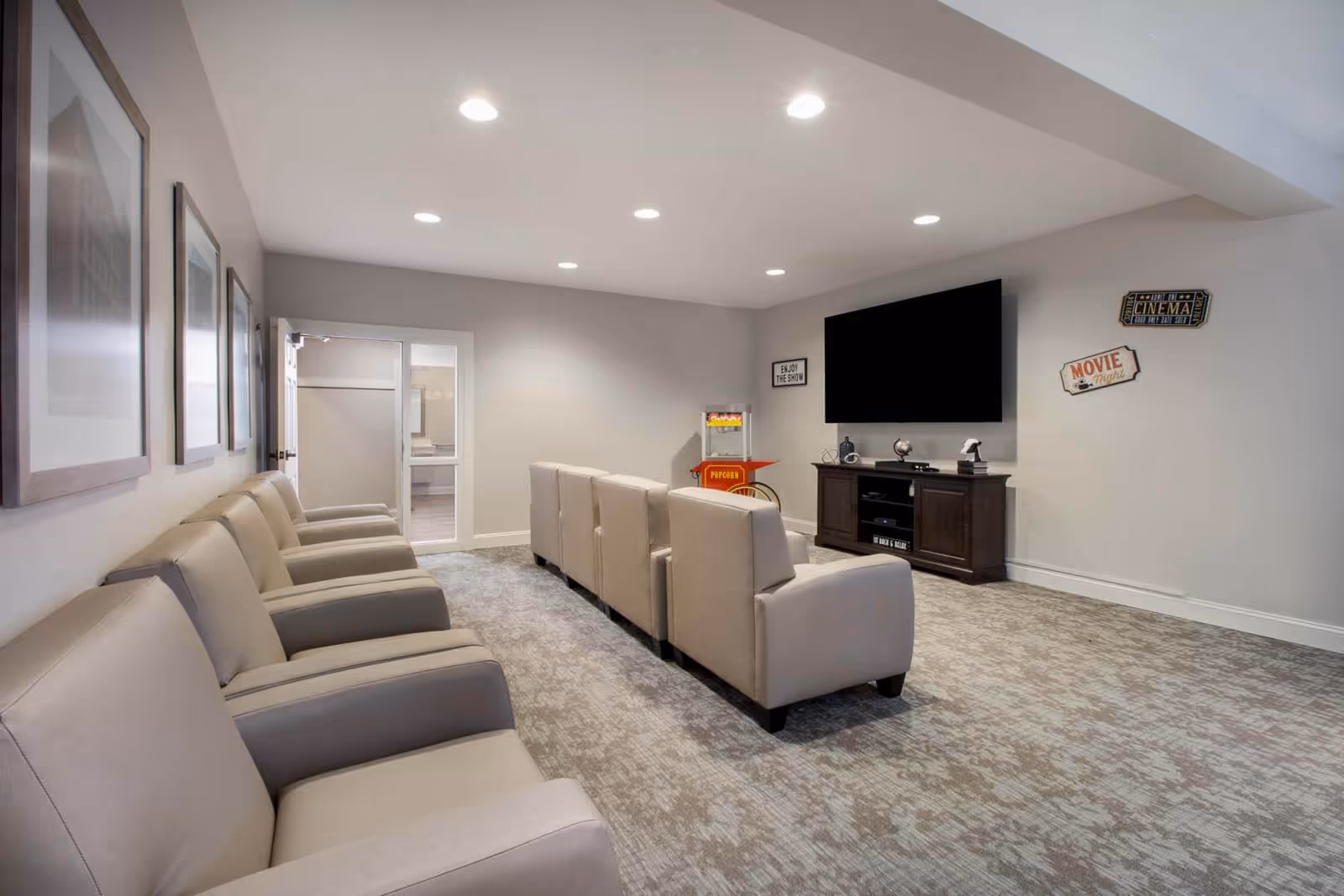Rows of beige armchairs facing a wall-mounted TV and cabinet in a small carpeted media/screening room with a popcorn machine.