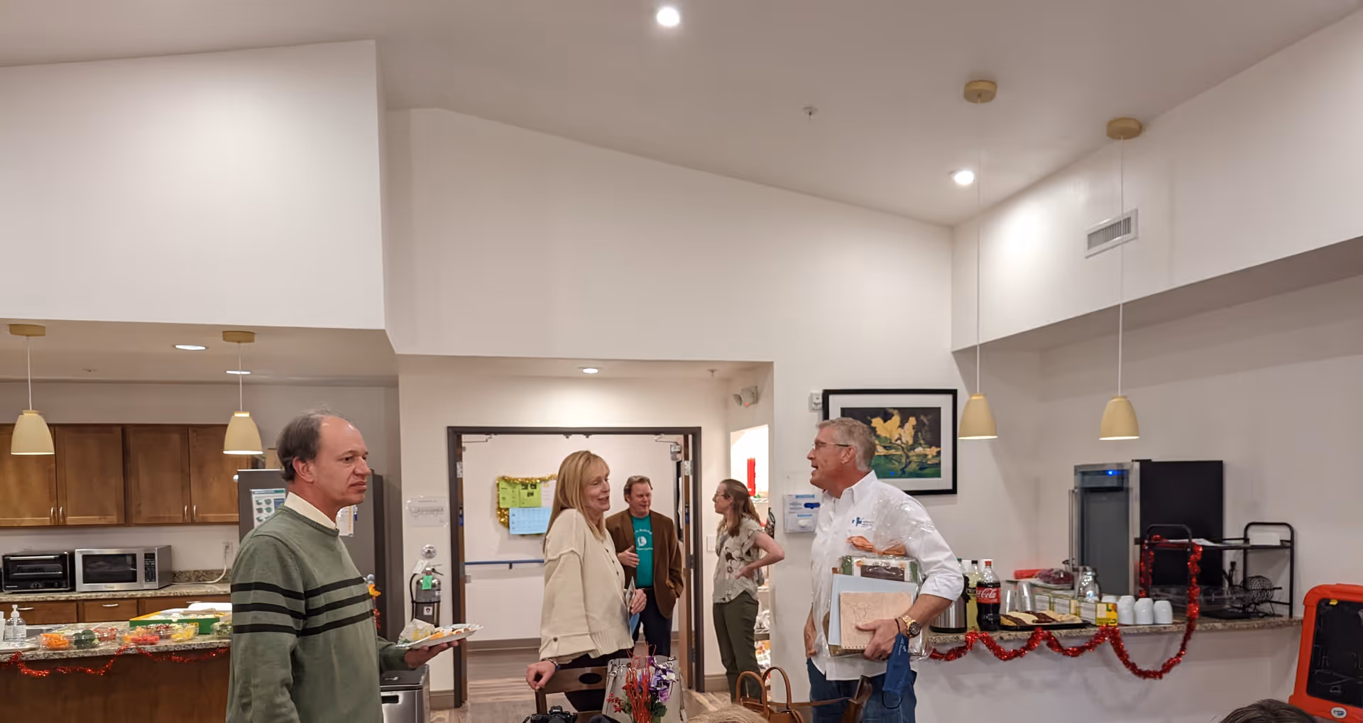 A group of five adults engaged in conversation in a well-lit common area with a kitchen counter in the background. The kitchen counter has various food items and drinks, and the room is decorated with red garland. The walls are white, and there are pendant lights hanging from the ceiling.