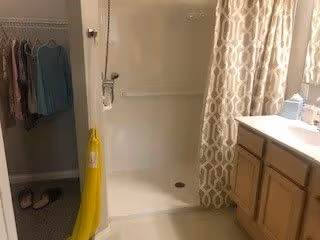 Small bathroom with a walk-in shower, patterned shower curtain, vanity with sink and cabinet, and an adjacent open closet with hanging clothes and slippers.