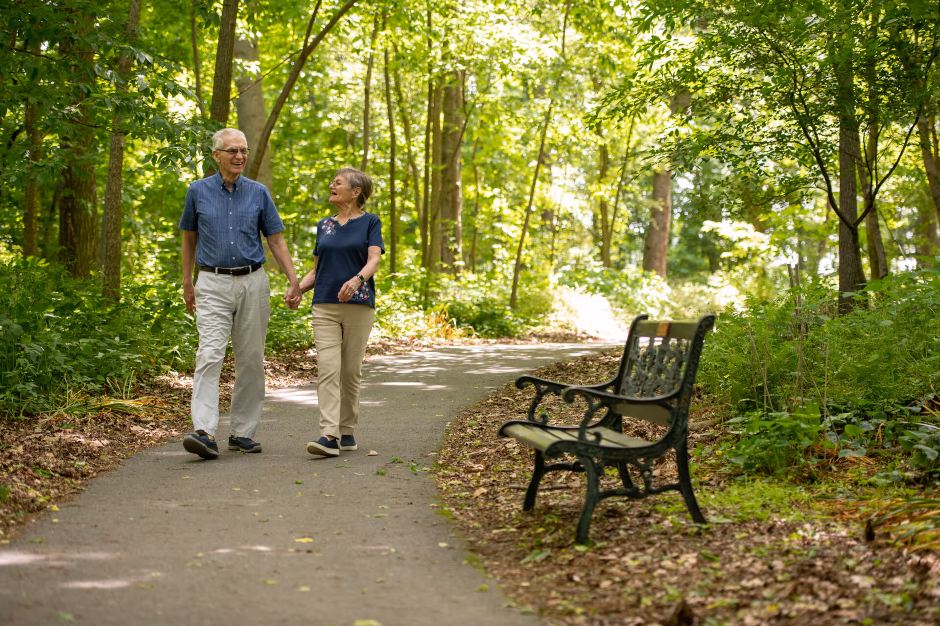 An elderly couple holding hands and walking along a paved path in a wooded area with lush green trees and foliage. There is a black metal bench with a wooden seat on the right side of the path.