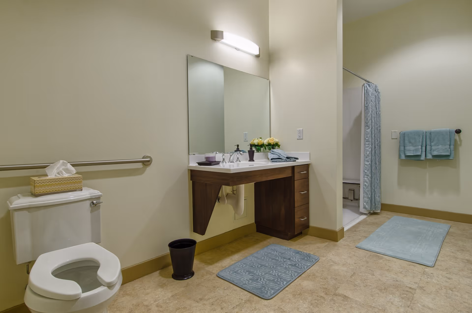 A clean and spacious bathroom with a white toilet that has a tissue box on top, a wall-mounted grab bar, a wooden vanity with a white sink and a large mirror above it, a small black trash bin, two blue bath mats on the floor, a shower area with a blue patterned curtain, and two blue towels hanging on a towel rack.