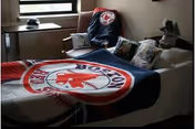A bed in a room covered with Boston Red Sox themed bedding including a blanket and pillows. There is a window with a small table underneath it, and a chair with a Boston Red Sox cover draped over it.
