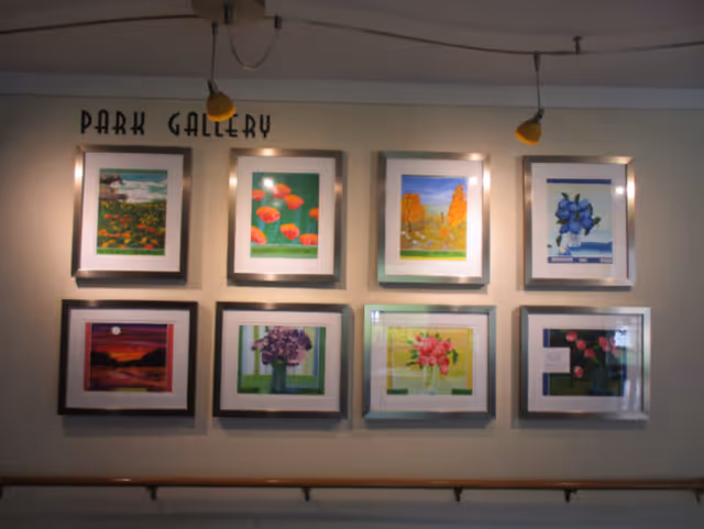 A wall display titled 'Park Gallery' featuring eight framed paintings of various flowers and nature scenes arranged in two rows of four. The paintings are illuminated by three overhead spotlights, and a wooden handrail runs along the bottom of the wall.