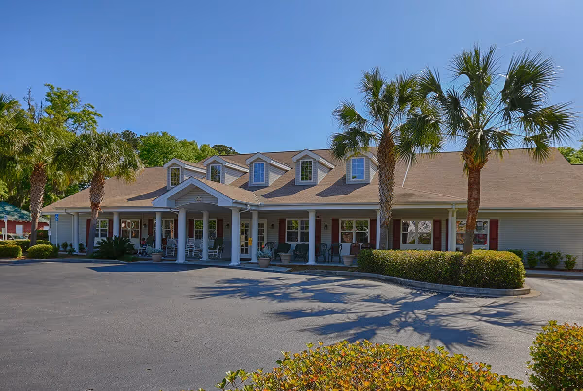 Front exterior of a single-story senior living facility with a covered porch, rocking chairs, and palm trees lining the driveway.