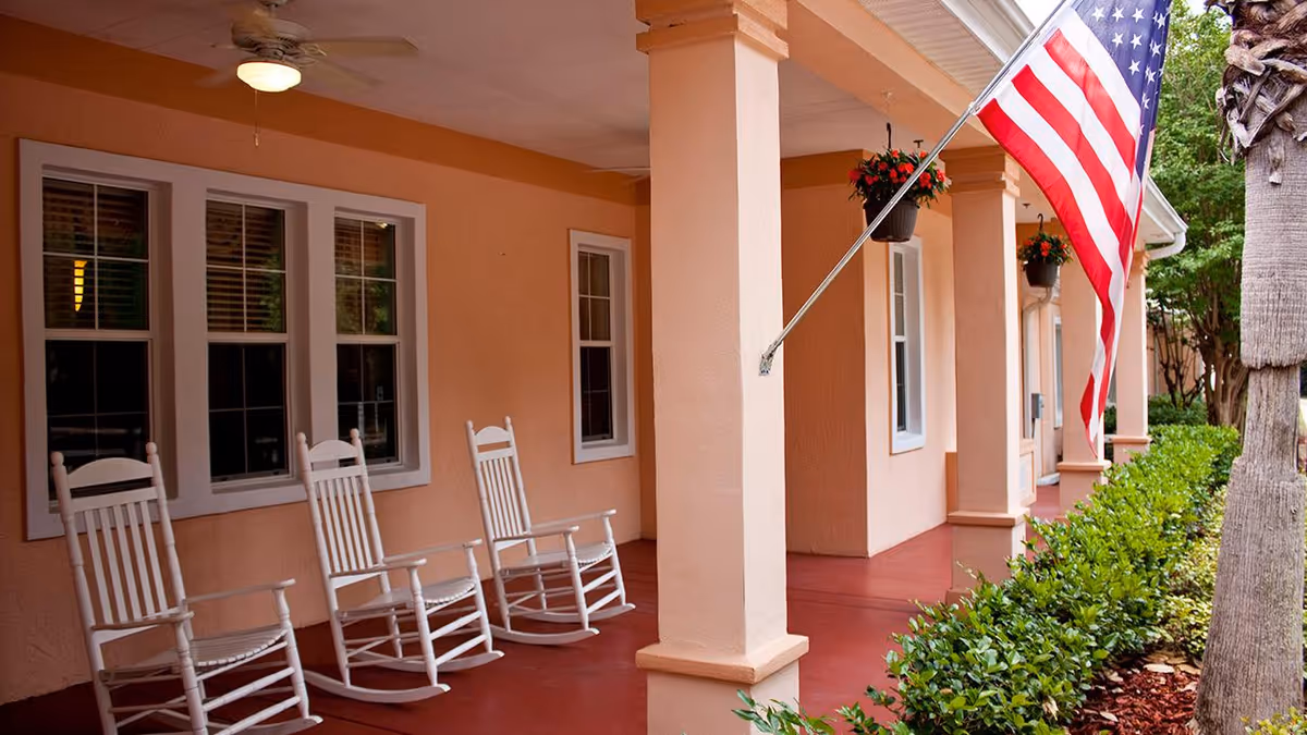 Covered porch area with three white rocking chairs lined up against a peach-colored wall with windows. There are columns supporting the roof, hanging flower pots, an American flag mounted on one of the columns, and green bushes along the edge of the porch.