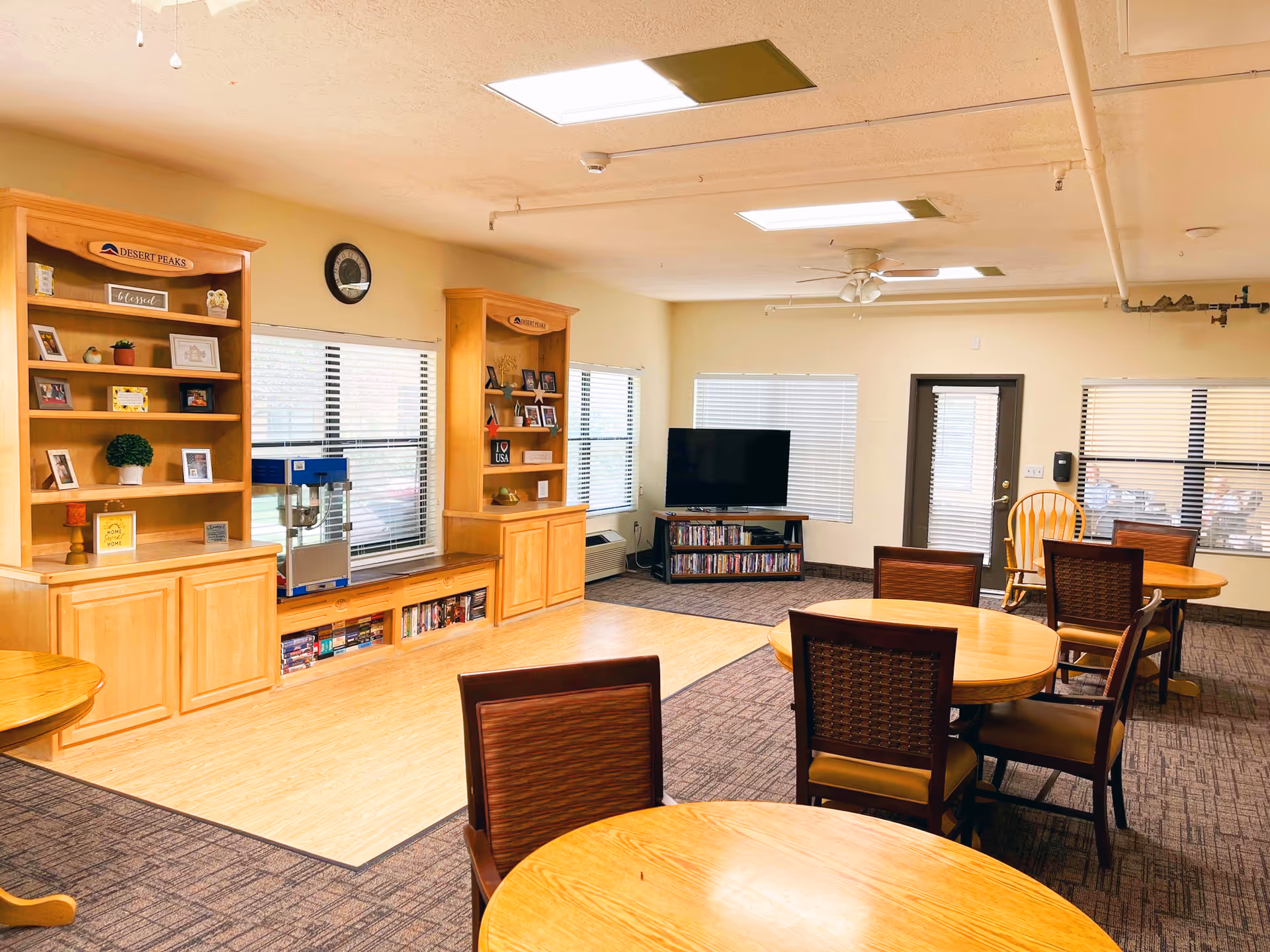 Bright communal dining and activity room with round wooden tables and chairs, built-in bookshelves, and a TV.