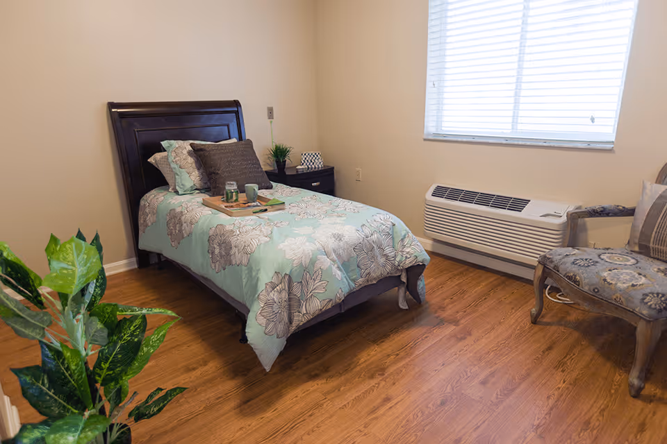 Bright single-occupancy bedroom with a floral-covered twin bed, nightstand, chair, window AC unit and wood flooring.